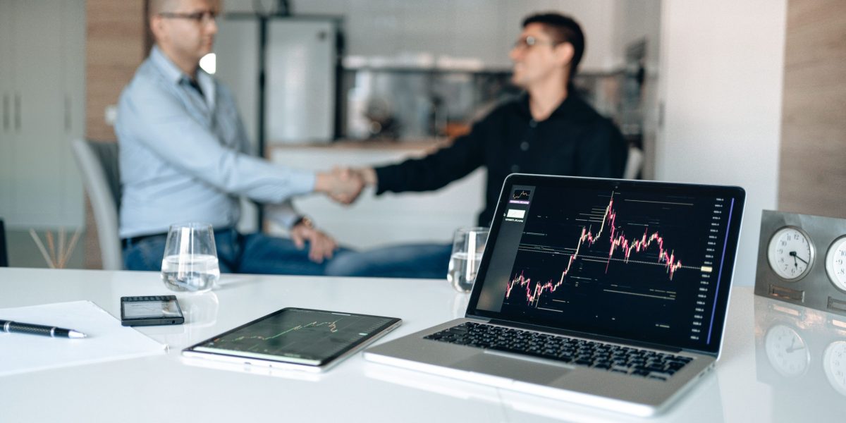 Two businessmen shake hands at a desk with stock market graphs on a laptop.